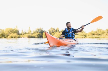 Can yeleği giyen bir adam sakin bir gölde kano sürüyor, güzel bir yaz gününde su sporlarının ve güneşin tadını çıkarıyor.