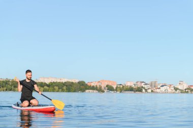 Genç sporcu, güneşli bir günde bir gölün arka planında bir şehir manzarasıyla ayakta duran bir kürek tahtasında kürek çekiyor.