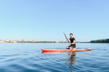 Genç sporcu kürek çekerken ayakta duran bir sörf tahtasında, sakin bir yaz gününün tadını çıkarırken doğa ile çevrili sakin bir gölde.