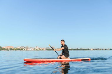 Genç sporcu, arka planda güneşli bir yaz gününün tadını çıkaran sakin bir gölde kürek çekiyor.