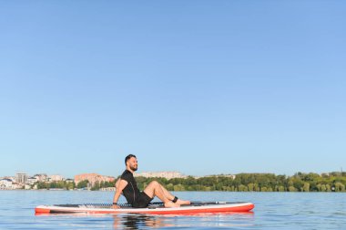 Genç sporcu yemek masasında oturmuş güneşli bir yaz gününde şehir nehrini seyrediyor ve eğleniyor.