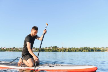 Genç adam yemek masasında diz çökmüş, güneşli bir yaz gününde sakin bir gölde kürek çekiyor, açık havayı kucaklıyor.