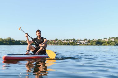 Genç adam ayakta duran bir kürek tahtasında dinleniyor, berrak mavi bir gökyüzünün altında sakin bir gölün sakin yaz atmosferinin tadını çıkarıyor.