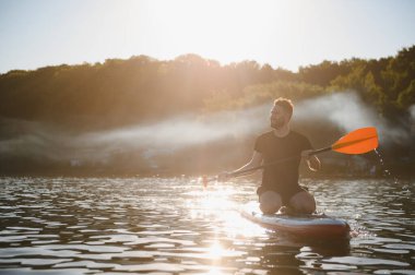 Genç adam gün batımında bir gölün üzerinde ayakta kürek çekiyor, altın saatin tadını çıkarıyor.