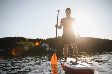 Genç adam gün batımında bir gölün üstünde kürek çekiyor, doğanın sükuneti ve güzelliğinin tadını çıkarıyor.