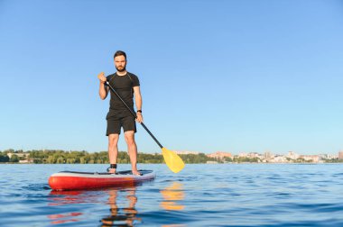 Genç sporcu yazın bir gölün kenarındaki yemek masasında kürek çekiyor. Suyun ve mavi gökyüzünün huzurunun tadını çıkarıyor.