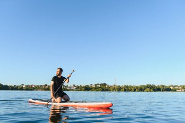 Genç adam ayakta duran bir kürek tahtasında güneşli bir yaz gününde sakin bir gölde kürek çekiyor ve boş zamanlarının tadını çıkarıyor.