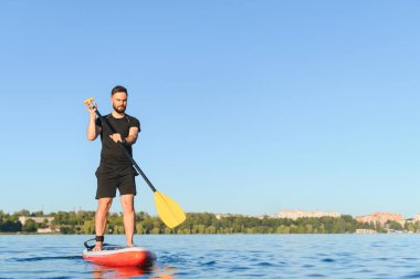 Genç sporcu kürek çekiyor, gölün üzerindeki bir panoda dengede duruyor, yazın su sporlarının tadını çıkarıyor.