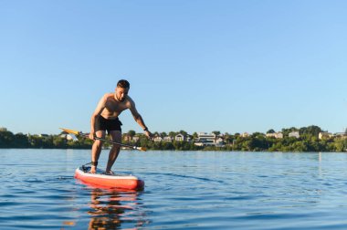 Genç atletik bir adam sakin bir gölde su sporunun ve tahtanın üzerinde dengede durmanın tadını çıkarıyor.
