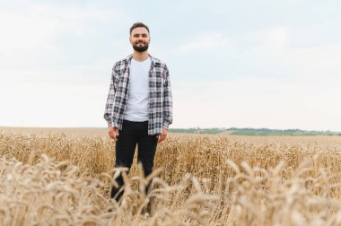 Genç çiftçi altın buğday tarlasında gururla duruyor, başarılı bir hasat mevsiminin meyvelerinin tadını çıkarıyor.