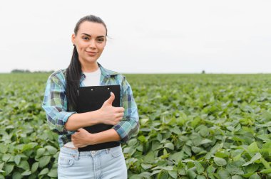 Kendine güvenen kadın tarım mühendisi, pano tutuyor ve yeşil bir soya tarlasında dikiliyor. Ürün büyümesinden duyduğu memnuniyeti dile getiriyor.