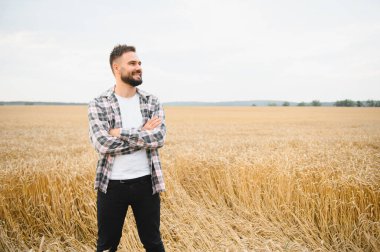 Altın buğday tarlasında kolları çapraz duran kendinden emin çiftçi, uzaklara bakarken ışıl ışıl gülümsüyor, gurur ve iyimserlik saçıyor.