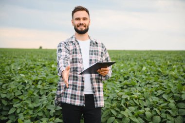 Tokalaşmak için elini uzatan tarımcı, pano tutan, yemyeşil soya tarlasında duran, ortaklığı ve tarım uzmanlığını sembolize eden.