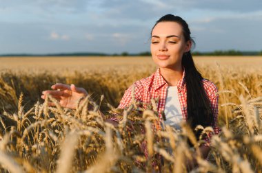 Genç kadın çiftçi gün batımında altın bir tarlada olgunlaşmış buğday kulaklarına hafifçe dokunarak emeğinin meyvelerinin tadını çıkarıyor.