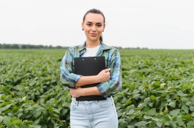Kendine güvenen kadın tarımcı elinde bir pano, yemyeşil soya bitkilerinin arasında duruyor, sağlıklı büyümeyi ve sürdürülebilir tarım uygulamalarını sağlıyorlar.