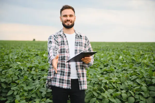 Tokalaşmak için elini uzatan tarımcı, pano tutan, yemyeşil soya tarlasında duran, ortaklığı ve tarım uzmanlığını sembolize eden.