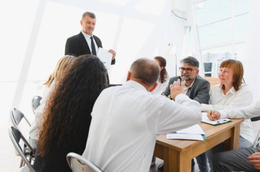 İş liderinin elinde belgeler var ve modern bir ofiste yaptığı toplantıda iş arkadaşlarına stratejiyi anlatıyor.