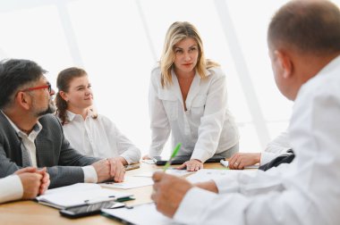 İş dünyası ekibi, şirket toplantısında mali raporları analiz ediyor ve stratejiyi tartışıyor. Kadın lider ayakta duruyor ve tartışmayı yönetiyor.