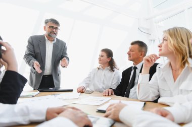 Deneyimli bir iş adamı modern bir ofiste bir şirket toplantısında ekibine seslenirken ayakta duruyor ve el kol hareketi yapıyor.