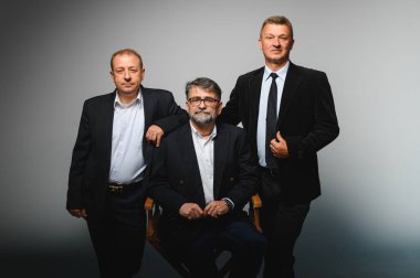 Üç işadamı bir stüdyoda poz veriyor, lider bir yönetmen koltuğunda oturuyor, güven ve takım çalışması gösteriyor.