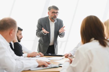 Olgun bir işadamı, parlak bir ofiste takım toplantısını yönetirken strateji ve planları tartışır.