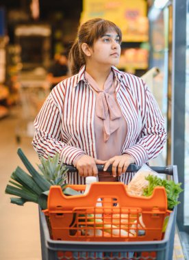 Genç Hintli kadın alışveriş arabasını iterken yoğun bir süpermarket koridorundan taze yiyecek seçiyor.