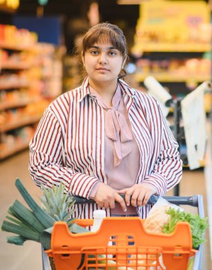 Kendine güvenen genç Hintli kadın marketin hareketli koridorlarında gezinirken içi taze yiyeceklerle dolu bir alışveriş arabasını itiyor.