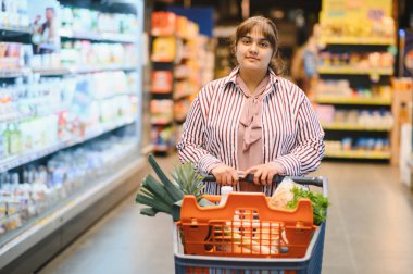 Genç Hintli bir kadın canlı bir süpermarketin hareketli koridorlarında gezinirken içi taze yiyeceklerle dolu bir alışveriş arabasını itiyor.