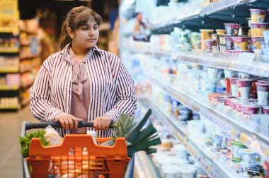 Genç Hintli kadın market koridorunda taze süt ürünleri seçerken alışveriş arabasını itiyor.