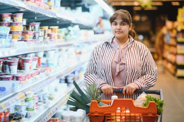 Genç Hintli kadın marketin hareketli koridorlarında gezinirken içi taze yiyeceklerle dolu bir alışveriş arabasını itiyor.
