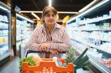 Genç Hintli bir kadın marketin koridorlarında gezinirken içi taze, sağlıklı yiyeceklerle dolu bir alışveriş arabasını itiyor.