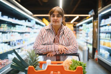 Genç Hintli kadın alışveriş arabasına yaslanıyor sağlıklı seçeneklerle dolu canlı bir süpermarket koridorunda taze erzakları karıştırıyor.