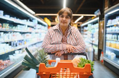 Genç Hintli kadın alışveriş arabasına yaslanıyor. Parlak, düzenli bir süpermarket koridorunda rahatça alışveriş yapıyor.