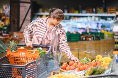 Sebze tezgahından domates seçen genç Hintli bir kadın markette taze ürünlerle dolu bir alışveriş arabasını itiyor.