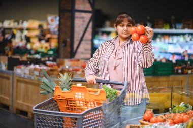 Müşteri, hareketli bir süpermarkette taze sebze alırken elinde canlı domates tutuyor.