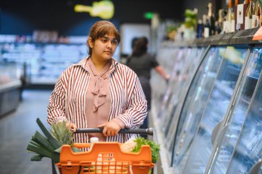 Genç Hintli kadın alışveriş arabasını itiyor ve dondurulmuş yiyecekleri marketin dondurulmuş bölümünden seçiyor.