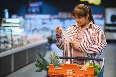Markette alışveriş arabasıyla duran ve makbuzu kontrol eden Hintli genç bir kadın.