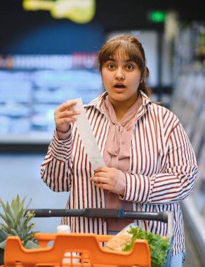 Genç Hintli kadın markette market fişini kontrol ederken şaşırmış görünüyor, yüksek fiyatlar ve enflasyondan duyduğu endişeyi dile getiriyor.