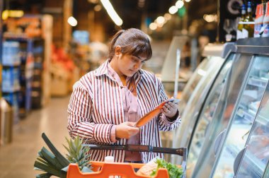 Alışveriş arabasını iten ve süpermarket buzdolabı koridorundaki donmuş gıda paketine etiket okuyan odaklanmış Hintli kadın.