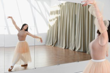 Genç bir kadın dans stüdyosunda bale figürleri yapıyor, etek ve tayt giyiyor, aynaya yansıyor.