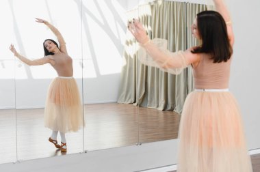 Şeftali rengi etekli genç bir kadın dans stüdyosunda büyük bir ayna ile bale hareketleri yapıyor.