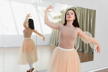 Genç balerin dans stüdyosunda bale hareketleri yapıyor, bej bir tayt ve tül etek giyiyor, zarafet ve kendini sanatına adamış.