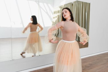 Genç balerin, bir dans stüdyosunda dans figürleri yapıyor, şeftali rengi etek ve tayt giyiyor, kararlı bir ifadeyle.