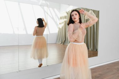 Genç balerin, dans stüdyosunda aynalarla dans ediyor şeftali rengi etek giyiyor ve kolları örgülü.