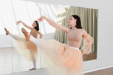 Genç bayan balerin dans stüdyosunda yüksek tekme pratiği yapıyor. Aynanın karşısında zarif bir etek giyiyor.