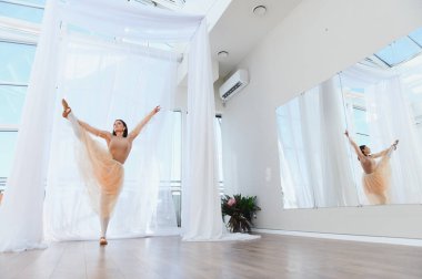 Genç bayan balerin bir dans stüdyosunda bale çalışıyor. Büyük camları ve aynaları var. Bir bacağını dikey olarak tutuyor.