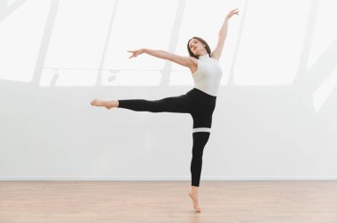 Modern bir bale pozu veren zarif dansçı, doğal ışıklandırılmış bir stüdyoda esneklik ve sanatçılık sergiliyor.