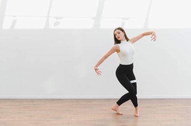 Dans öğrencisi, ahşap zemini ve beyaz duvarları olan parlak bir stüdyoda modern bir dans koreografisi uyguluyor.