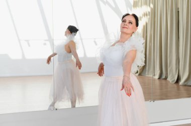Yaşlı balerin dans stüdyosunda dans figürleri yapıyor. Beyaz tayt ve pembe etek giyiyor. Aynaya yansıyor.