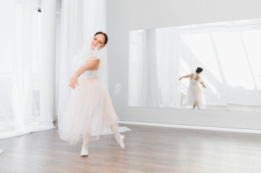 Yaşlı balerin dans stüdyosunda dans figürleri yapıyor. Aynalı, beyaz etekli.